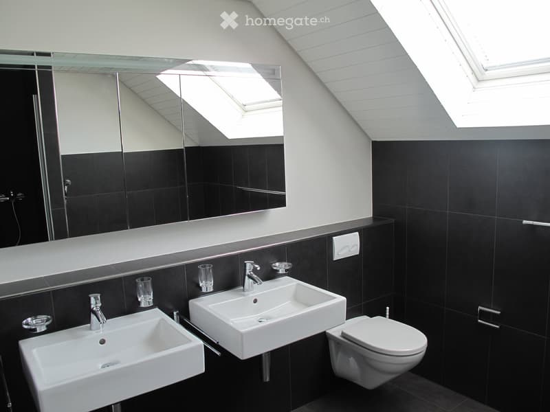 Modern bathroom with black tiled walls, two white sinks, a toilet, a large mirror, and two glass cups on the counter.