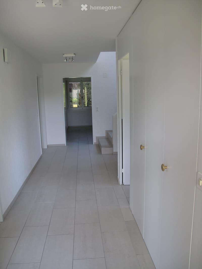 Empty hallway with tiled floor, white walls, and recessed lighting. Features a small step leading to an open doorway with blinds.