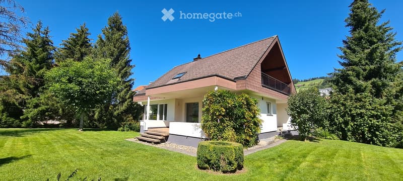Two-story house with a red-tiled roof, white walls, and a balcony on the upper level, surrounded by a well-maintained lawn and tall trees.