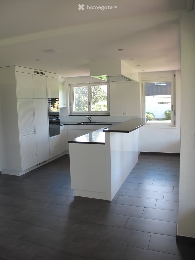 Modern kitchen with white cabinets, dark countertops, and a central island. Features include a sink, oven, and large windows providing natural light.