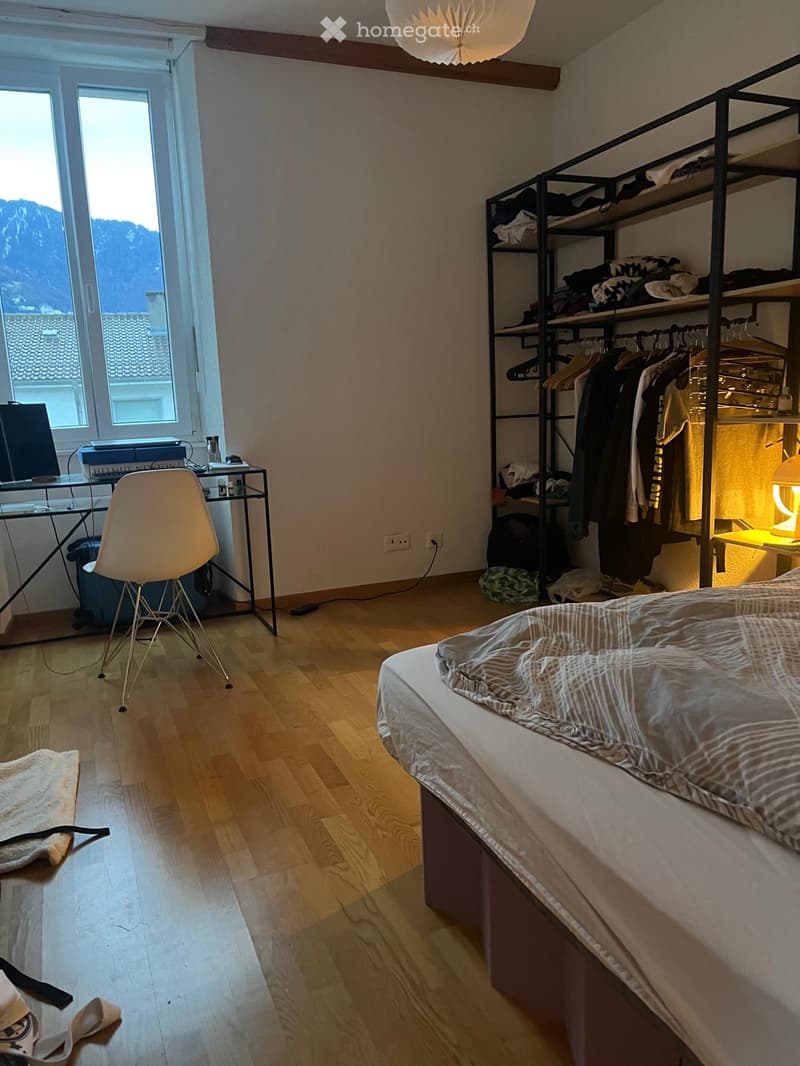 Cozy bedroom with wooden flooring, a neatly made bed, a modern chair, a desk with a computer, and a clothing shelf with hanging clothes.
