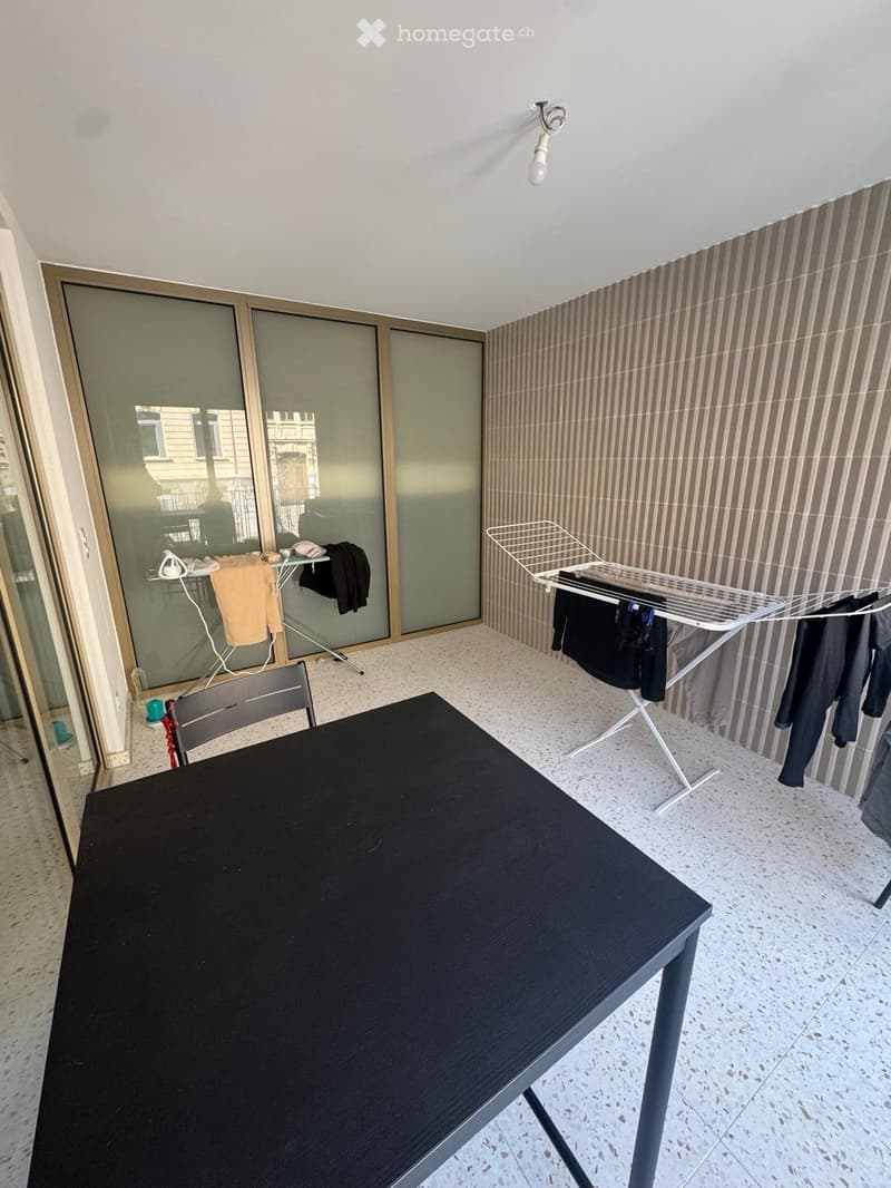 Balcony with a black table, a chair, an ironing board, and a clothes drying rack with hanging clothes.