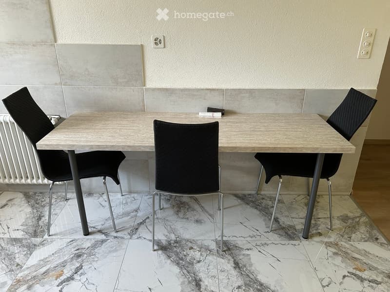 Dining area with a light wood table and four black chairs on a marble floor.