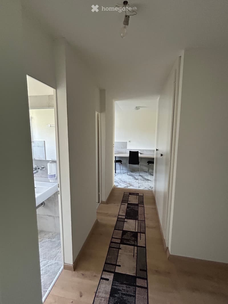 A narrow hallway with beige walls and a patterned runner rug, leading to a room with a desk and chairs.