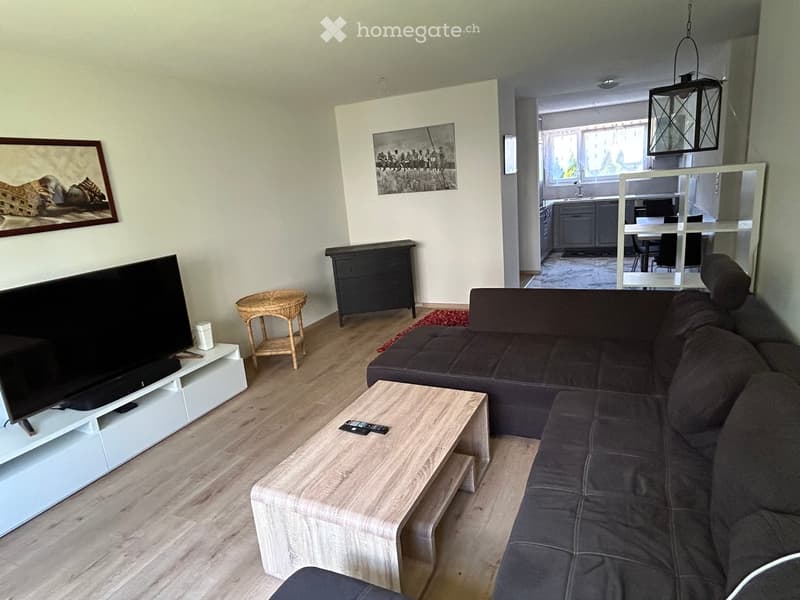 Modern living room with a dark grey sectional sofa, light wooden coffee table, white TV stand, wicker side table, and a small cabinet. The room features light-colored walls, wooden flooring, and artwork on the walls.