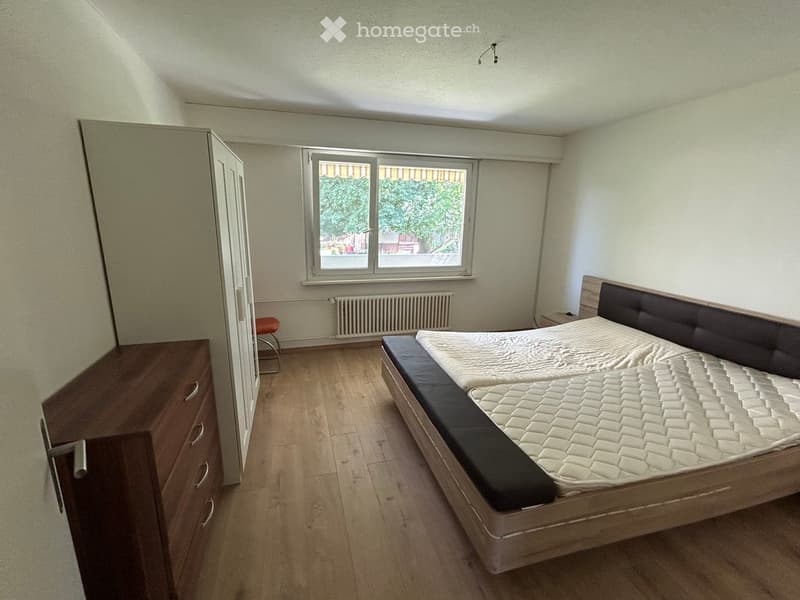 Bedroom with a double bed, wardrobe, and a small stool near the window. The room has wooden flooring and white walls.
