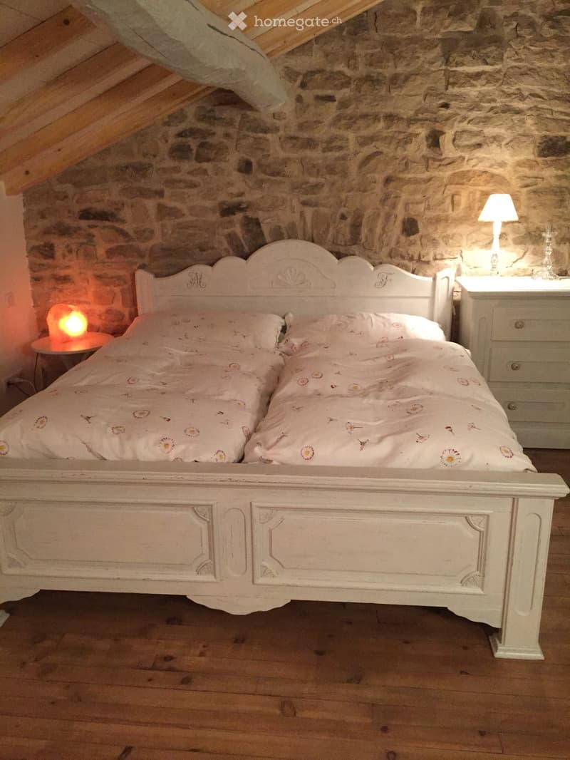 Cozy attic bedroom with exposed wooden beams and stone walls, featuring a white wooden bed with floral bedding, a nightstand with a lamp, and a small table lamp on the floor.