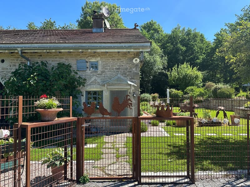 A rustic stone house with a metal gate, surrounded by a well-maintained garden with potted plants, decorative metal roosters, and lush greenery.