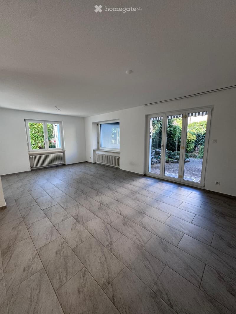 Empty room with tiled floor, white walls, large windows, and a sliding glass door leading to an outdoor area.