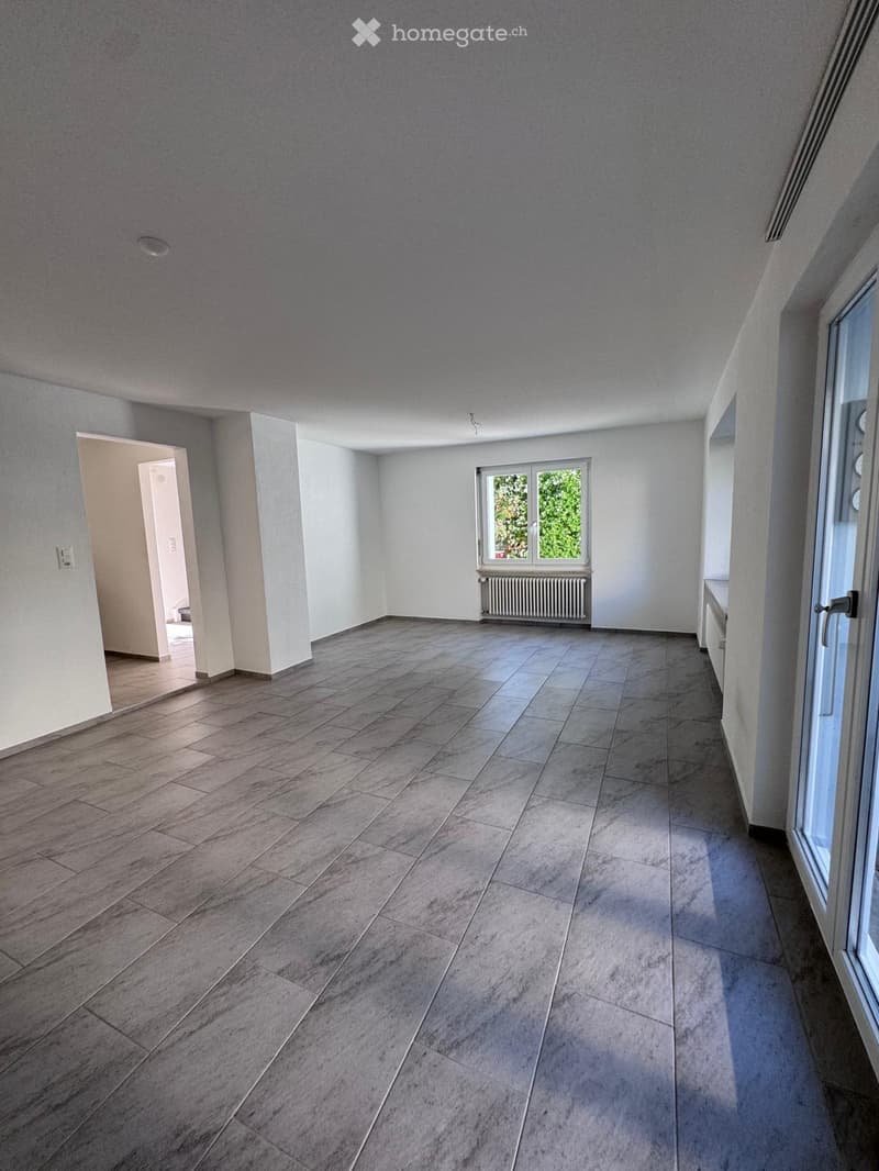 Empty room with light grey walls, tiled floor, and large windows allowing natural light. There is a radiator under one window and a doorway leading to another room.