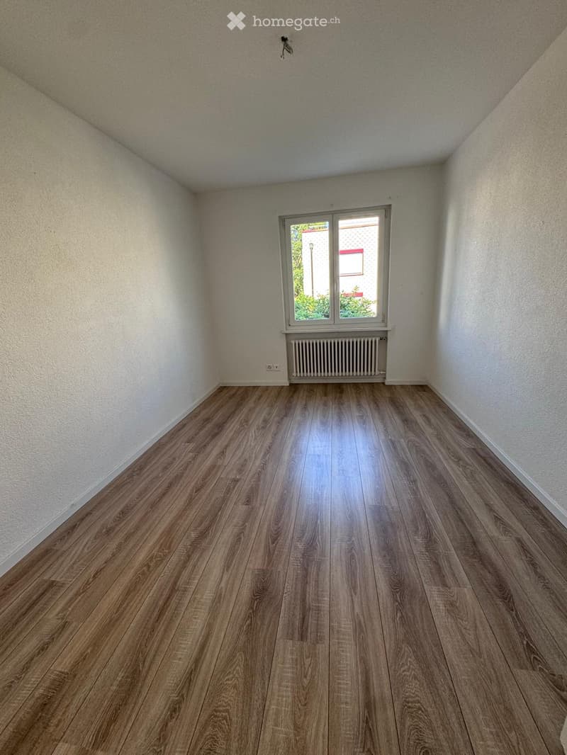 Empty room with wooden flooring, white walls, and a window with a radiator below it.