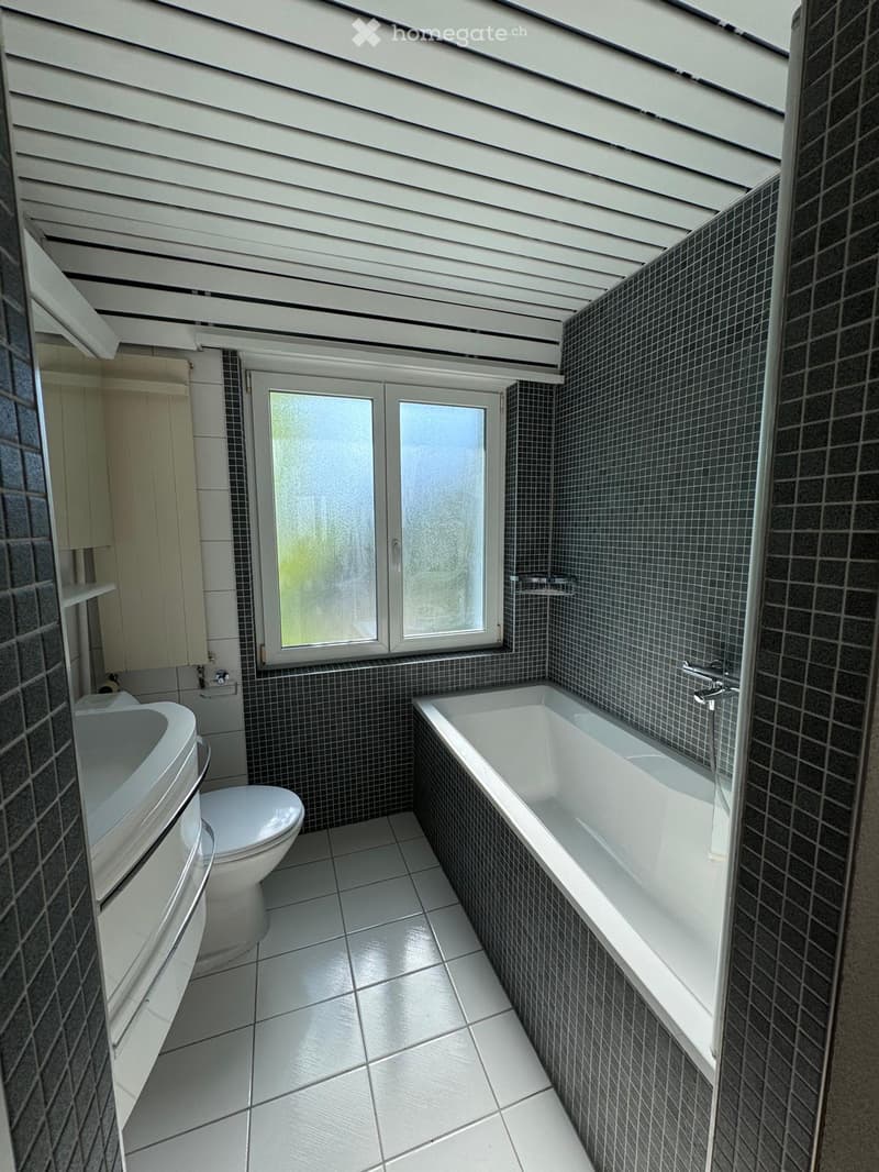 Modern bathroom with a bathtub, toilet, and sink, featuring dark tiled walls and a large window with frosted glass.