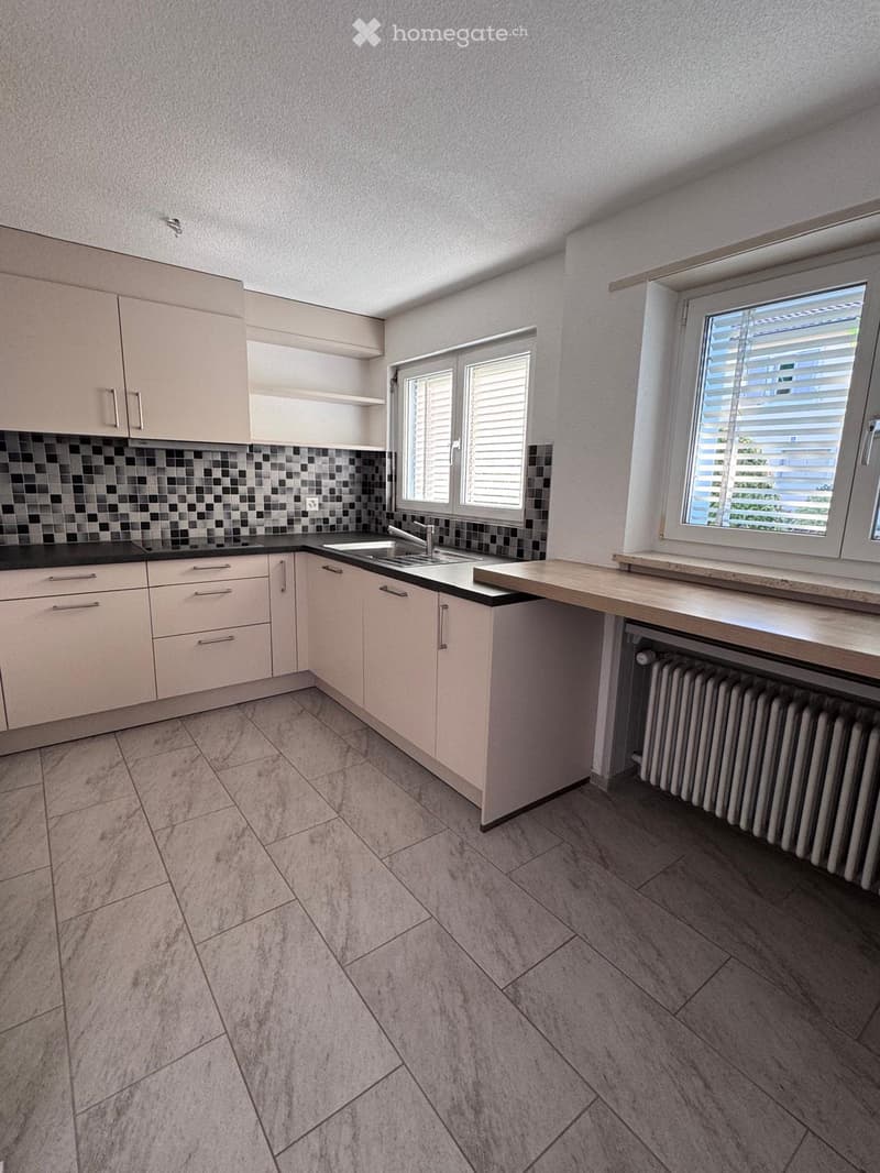 Empty kitchen with light-colored cabinets, a sink, and tiled backsplash. The space includes a countertop, windows, and tiled flooring.