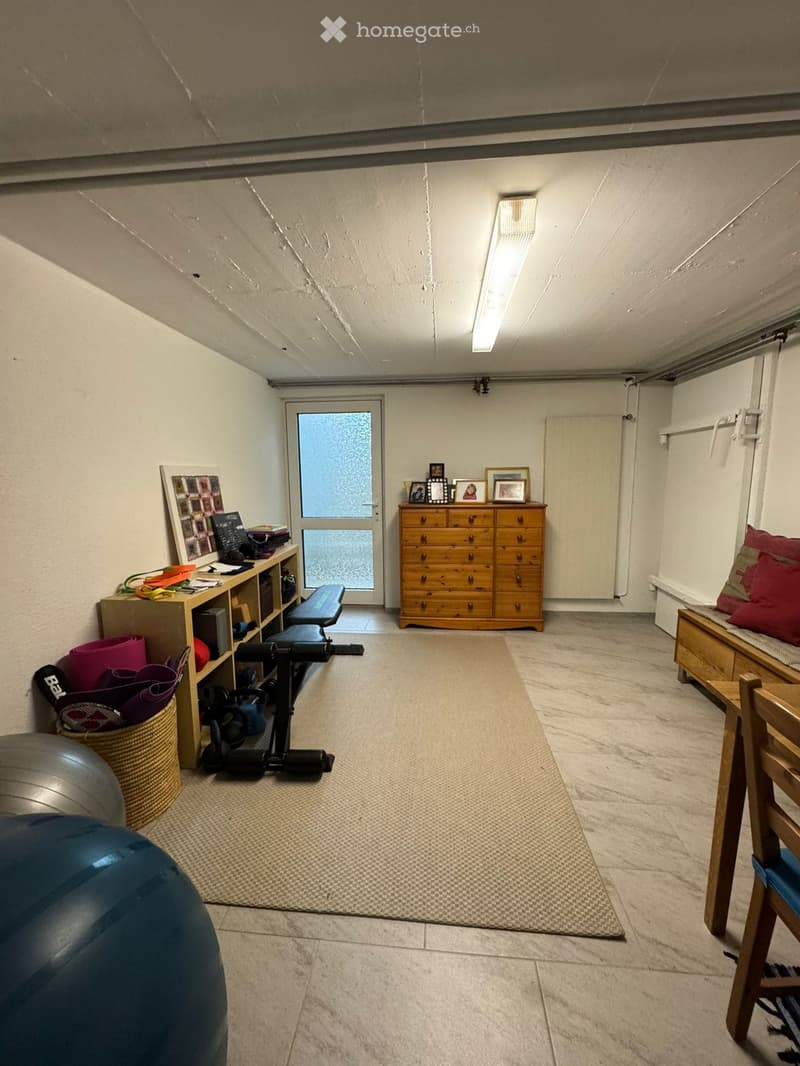 The room is a well-lit, furnished space with a mix of furniture and fitness equipment. It features a wooden shelf with various items, a large wooden drawer unit with picture frames on top, a small bench with pillows, a yoga mat, a fitness bench, a fitness