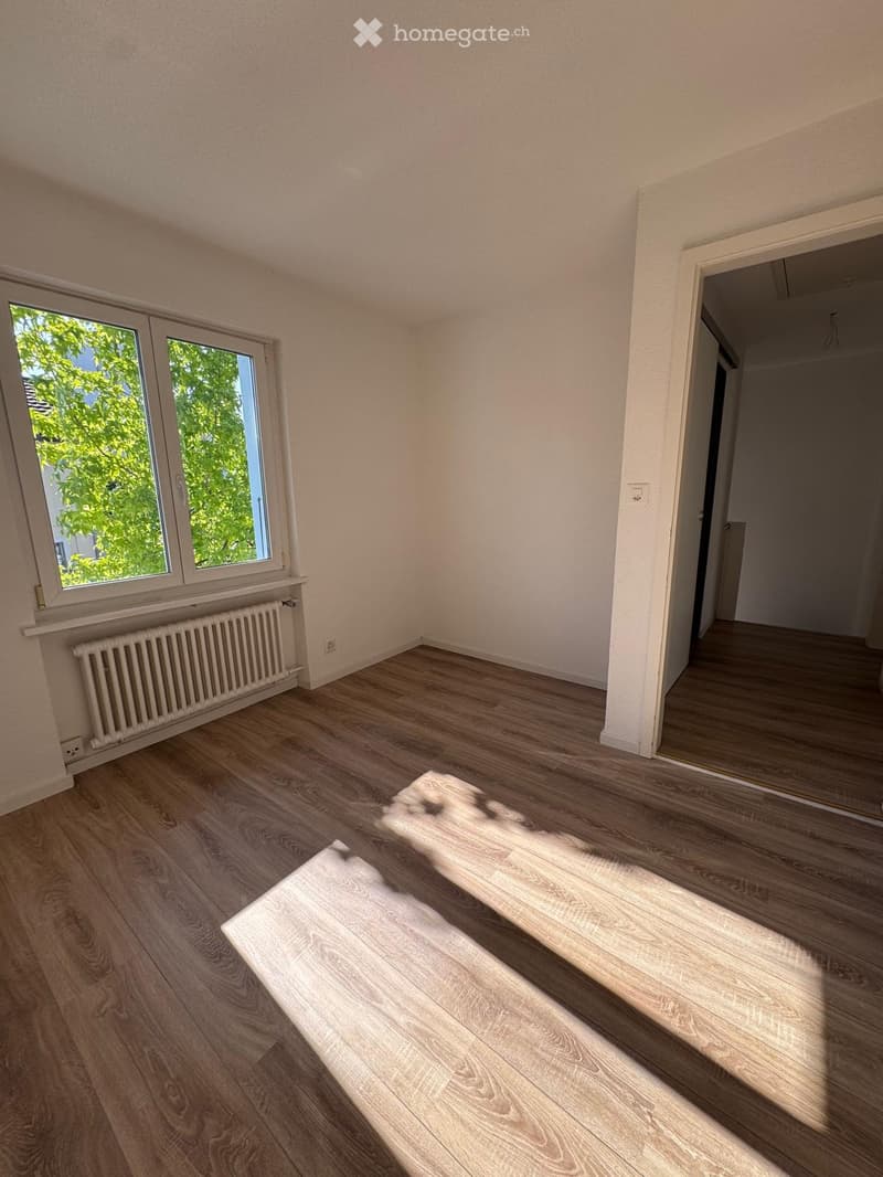 Empty room with light wooden flooring, white walls, and a window with a radiator below it. The room has a doorway leading to another space.