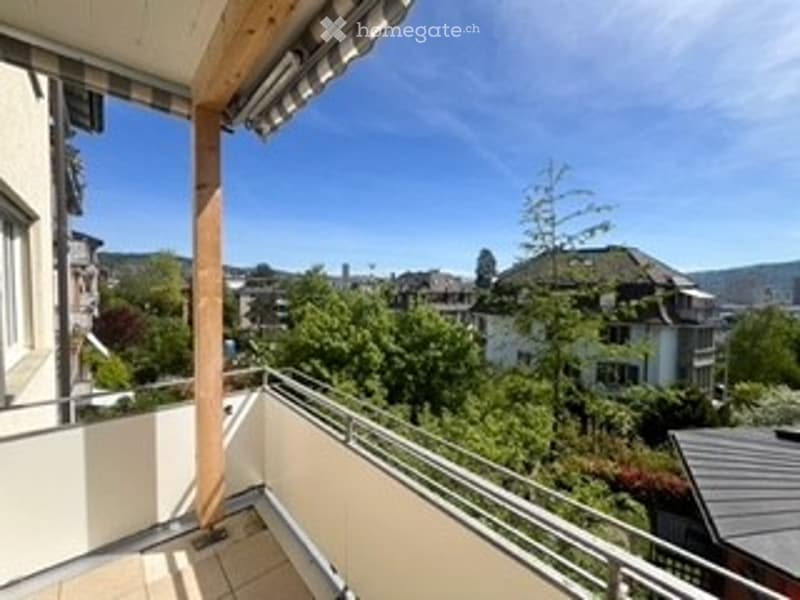 Balcony with wooden railings, overlooking a green, tree-filled neighborhood with houses and a clear blue sky.