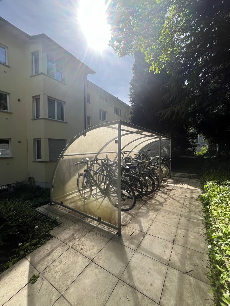 Outdoor area with tiled floor, covered bicycle shelter, several bicycles, and surrounding greenery.