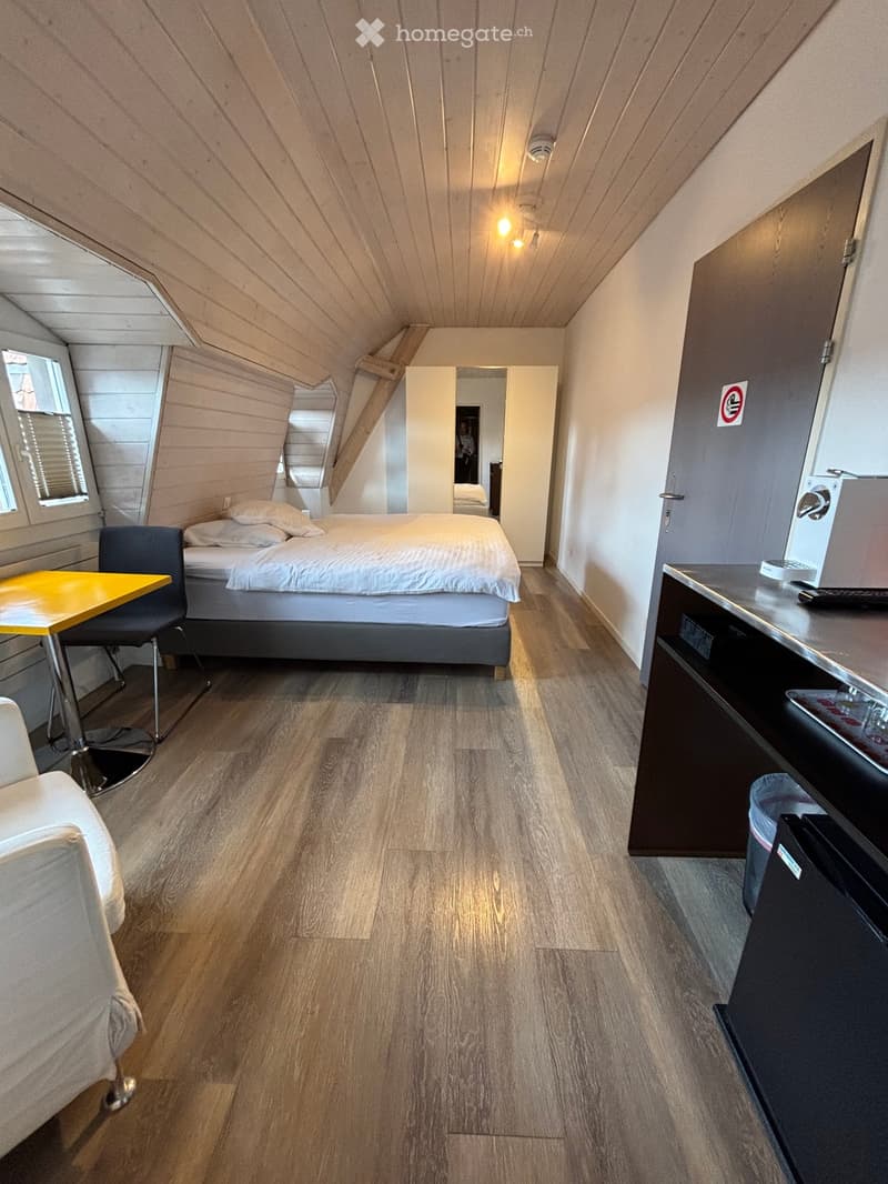 Cozy attic bedroom with wooden floors, slanted wooden ceiling, bed, chair, small table, and a mini-fridge.