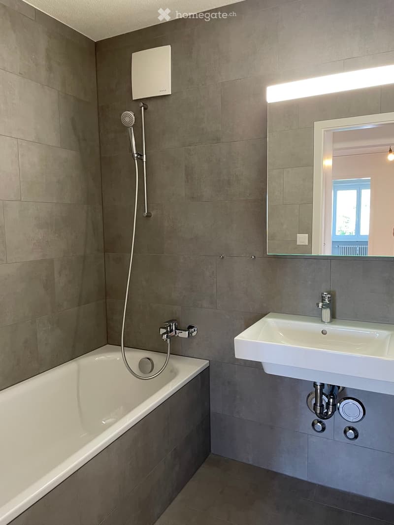 Modern bathroom with a bathtub, sink, mirror, and showerhead. Grey tiled walls and floor, white fixtures.