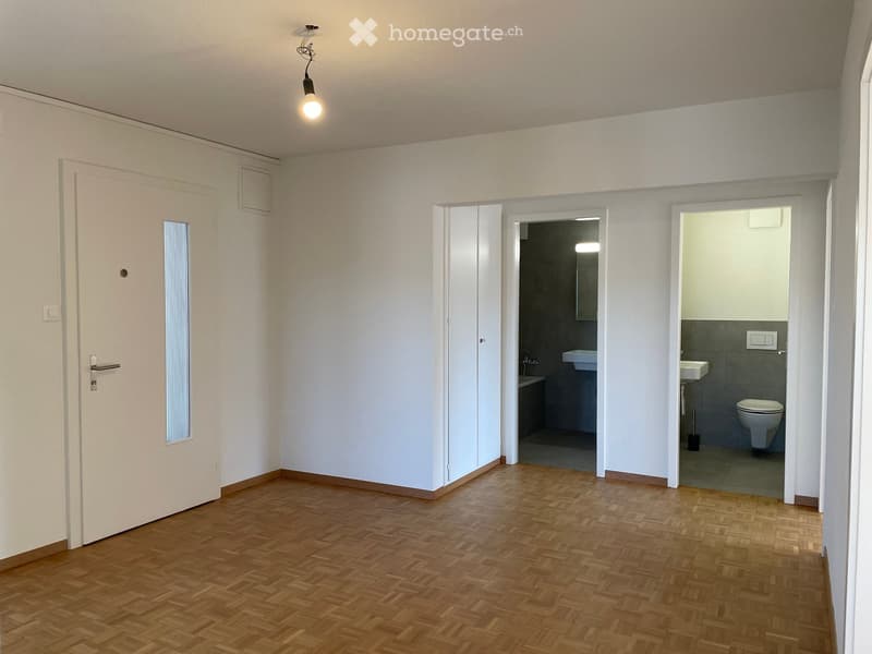 Empty room with wooden herringbone flooring, white walls, and two adjacent bathrooms visible through open doorways.