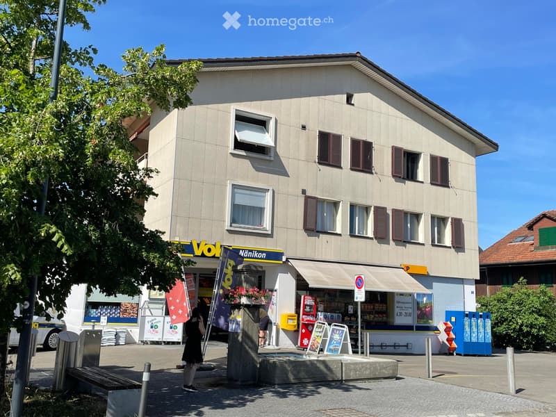 Two-story commercial building with a storefront labeled "Vola Nänikon" and a small awning. Windows with shutters, signage, and a few people near the entrance.