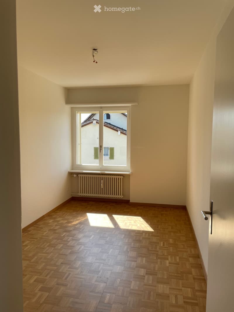Empty room with wooden herringbone flooring, white walls, and a large window with white frame and radiator below. No furniture or equipment present.