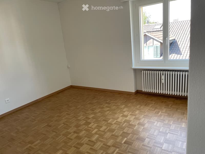 Empty room with wooden herringbone flooring, white walls, and a window with a radiator below it.