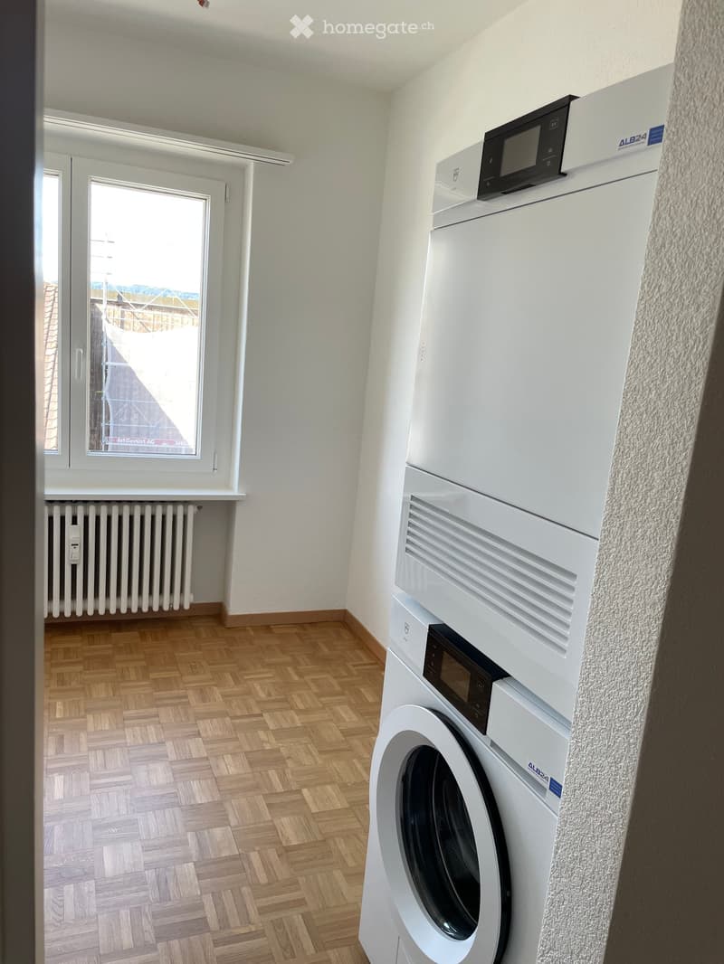 A small room with wooden flooring, a white washing machine, and a radiator under a window.