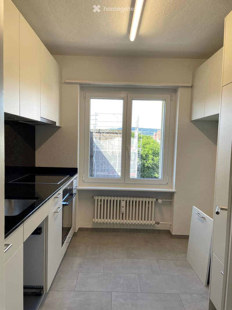 Empty kitchen with white cabinets, black countertop, built-in oven, radiator, and large windows overlooking a view.