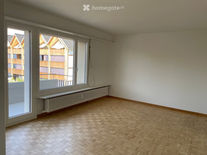 Empty room with wooden herringbone flooring, white walls, large windows, radiator, and a sliding door leading to a balcony.
