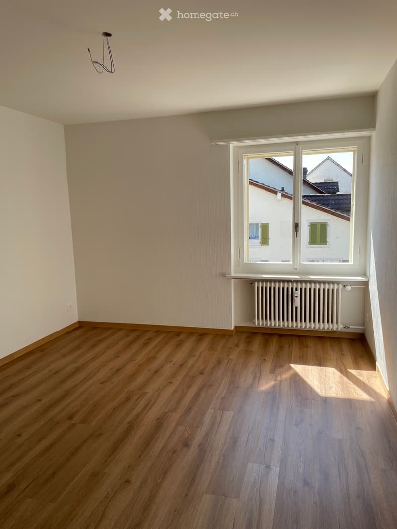 Empty room with wooden flooring, white walls, and a large window with a radiator below it.