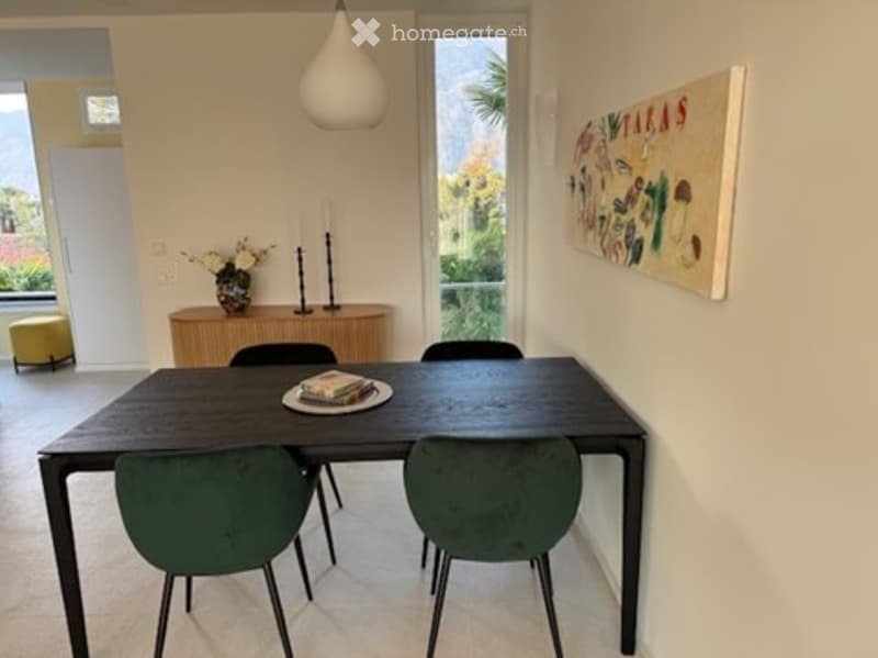 Dining room with a dark wooden dining table, four green upholstered chairs, a sideboard with a vase and candle, a yellow stool, and a colorful painting on the wall.