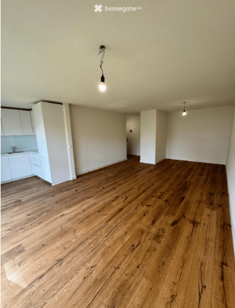 Empty room with wooden flooring, white walls, and a small kitchen area visible in the background.