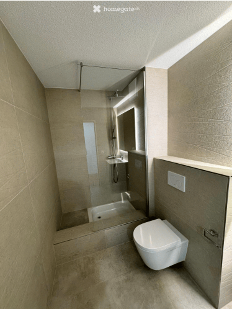 Modern, minimalist bathroom with light beige tiles, a toilet, a shower area with glass enclosure, and a sink.