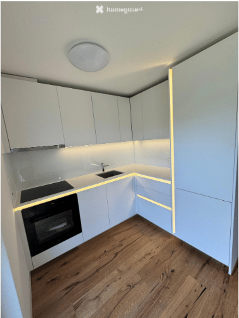 Modern, minimalist kitchen with white cabinets, wooden flooring, and built-in appliances including an oven and sink.