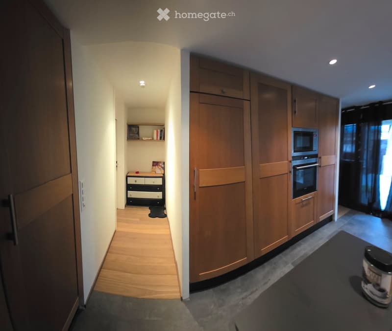 A narrow hallway with wooden flooring and white walls, leading to a small room with a dresser and a kitchen area with wooden cabinets and built-in ovens.