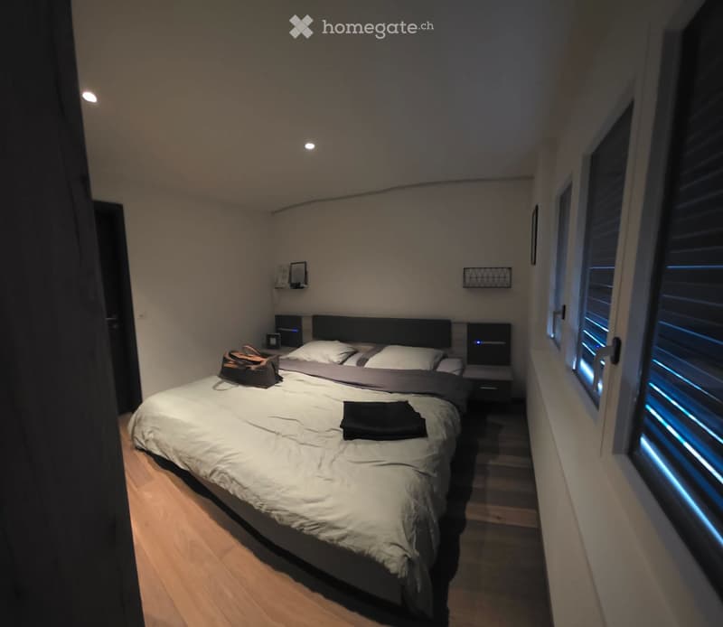 Modern bedroom with a large bed, nightstands, and a handbag on the bed. The room features wooden flooring and large windows with blinds.