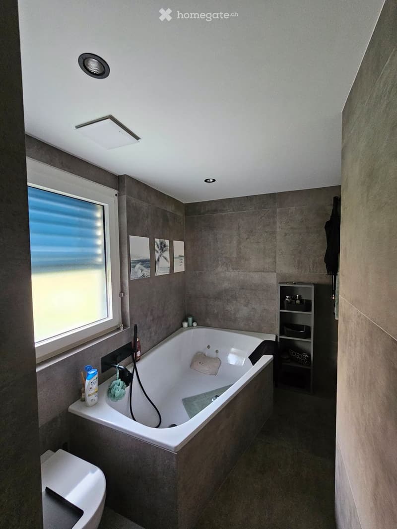 Modern bathroom with a large bathtub, toilet, shelf, and window offering a scenic view. The room features grey tiles, a sleek design, and decorative picture frames on the wall.