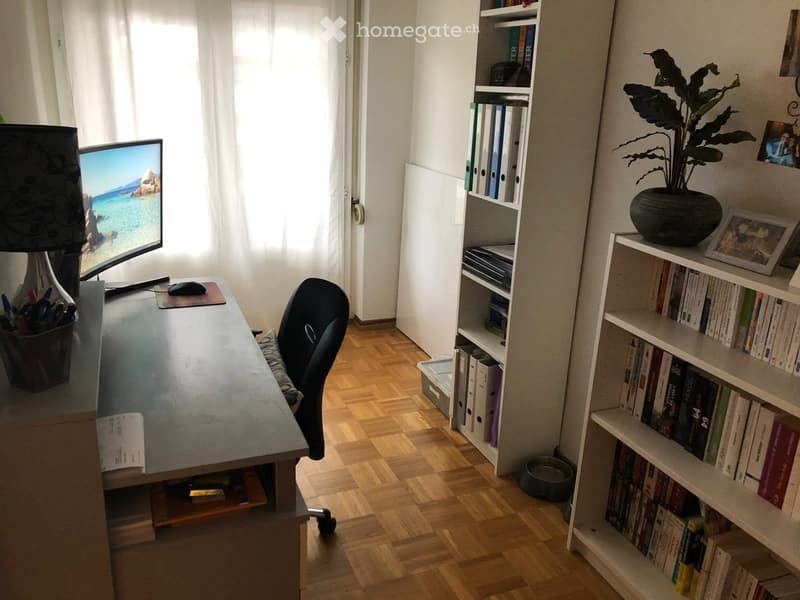 A well-lit study or home office with a wooden floor, a large desk with a computer screen, a chair, a lamp, shelves filled with books, and a potted plant on a shelf.