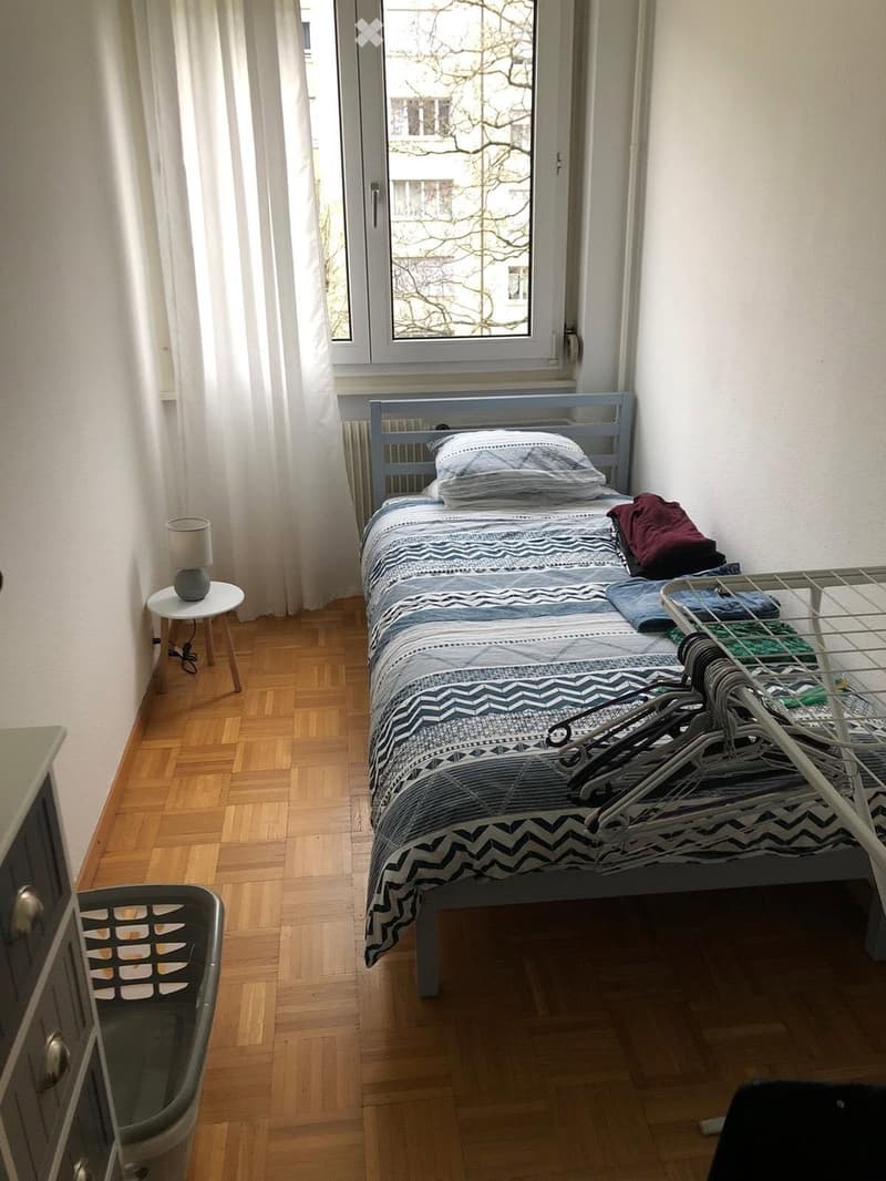 A small bedroom with a single bed, a bedside table with a lamp, a radiator, and a wardrobe. The bed is made with a patterned duvet and pillows, and there are some folded clothes on the bed.