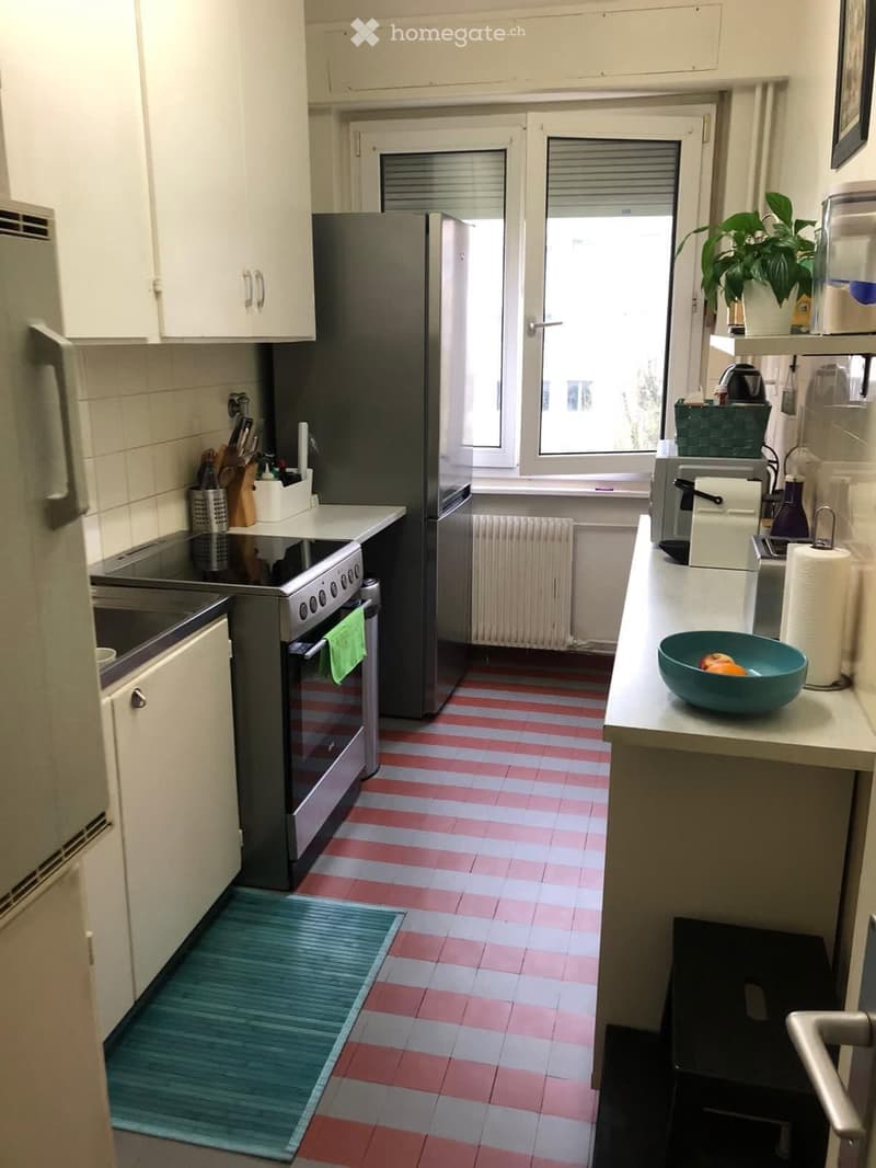 A compact kitchen with white cabinets, a stainless steel refrigerator, an oven, a sink, and a countertop with a bowl of fruit. The floor features a red and white tiled pattern.