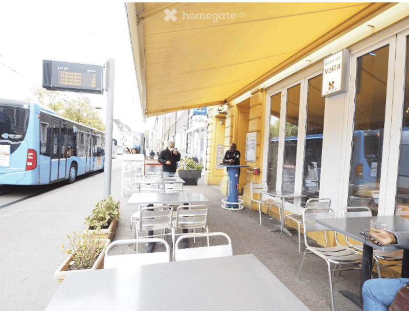 Outdoor seating area of a café with tables and chairs, yellow awning, potted plants, and a bus passing by on the street.