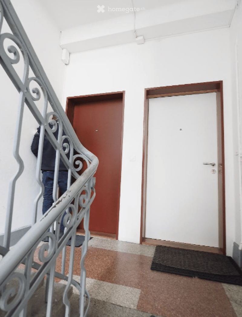 Hallway with white walls, a red door, a white door, a black doormat, and a decorative metal railing leading upstairs.
