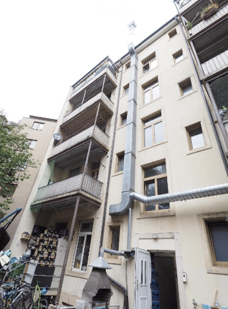 Multi-story residential building with balconies, windows, and external pipes. The ground floor has a doorway and some stacked items near the entrance.