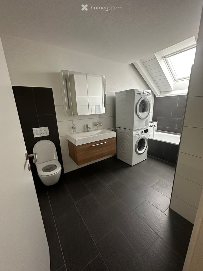Modern bathroom with dark tiled floor, white sink with wooden cabinet, toilet, mirror, washing machine, and bathtub.