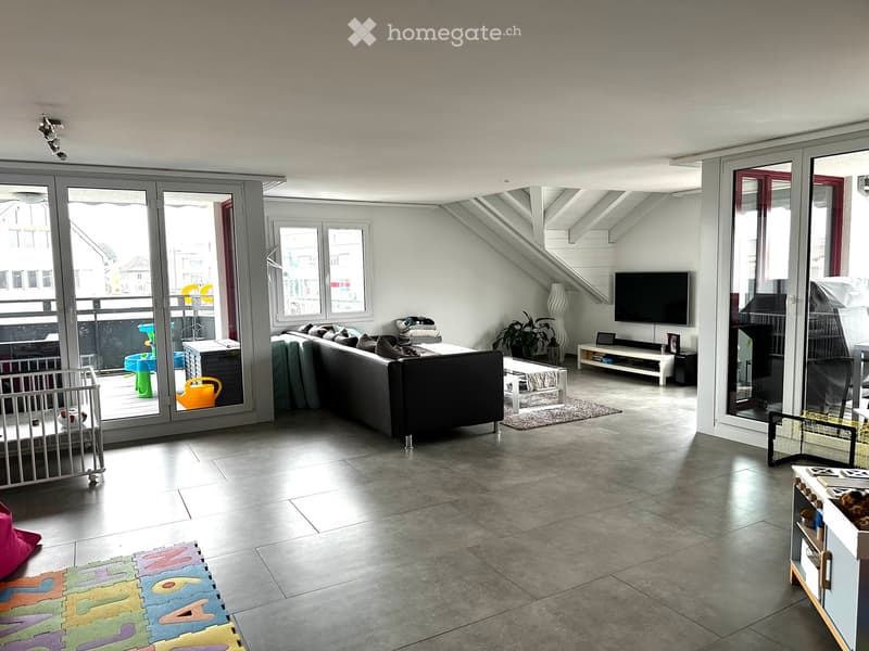 Modern living room with large windows and sliding glass doors leading to a balcony. Features a grey sectional sofa, coffee table, TV stand, and decorative plant. Includes a baby gate and colorful play mat on the floor.