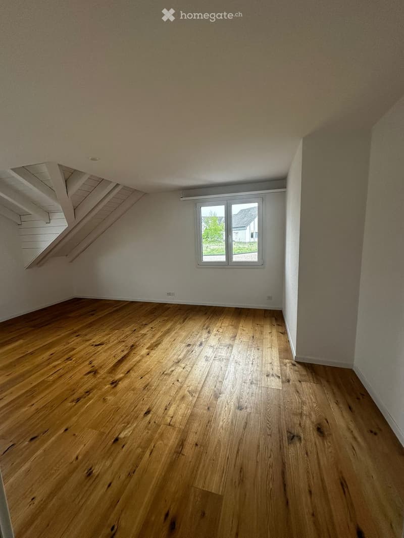 Empty room with wooden flooring, white walls, and a window with a view of the outside. The ceiling has exposed wooden beams and a skylight.