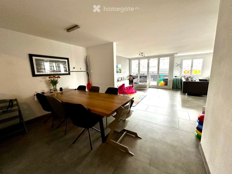 Open-plan living and dining area with a wooden dining table, chairs, a sofa, and decorative items including a vase and picture frame.