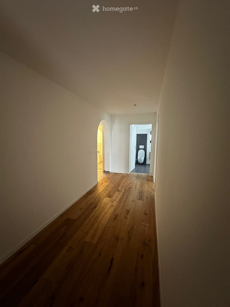 Empty hallway with wooden flooring and white walls, leading to a bathroom with a toilet and sink visible at the end.