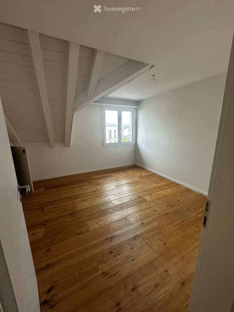 Empty attic room with wooden flooring, white walls, and a single window letting in natural light. The ceiling has exposed wooden beams and a skylight.